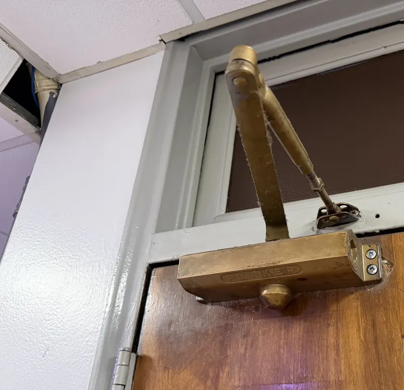 Brass hydraulic overhead door closer with parallel arm mounted on a wooden door below a transom