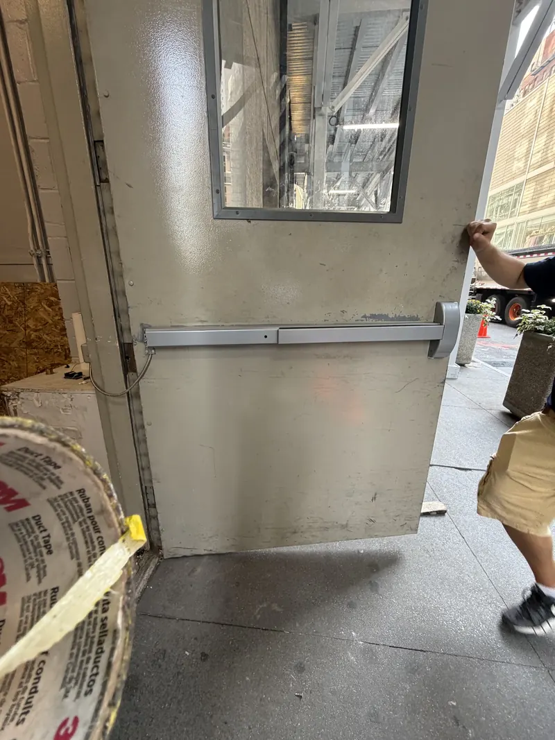 Gray commercial steel door with a horizontal panic bar exit device, being held open by a technician