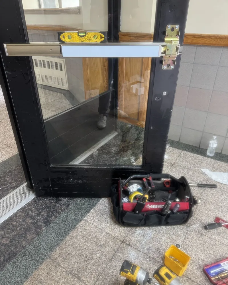 Spirit level resting on a new exit device being installed on a black aluminum entrance door, with tools on the floor