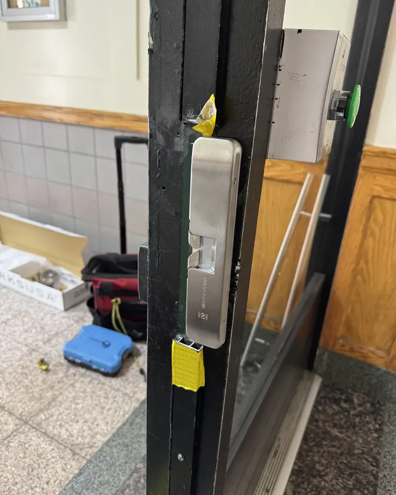 Stainless steel faceplate and latch of a mortise lock set into the edge of a black storefront door, with tools on the lobby floor