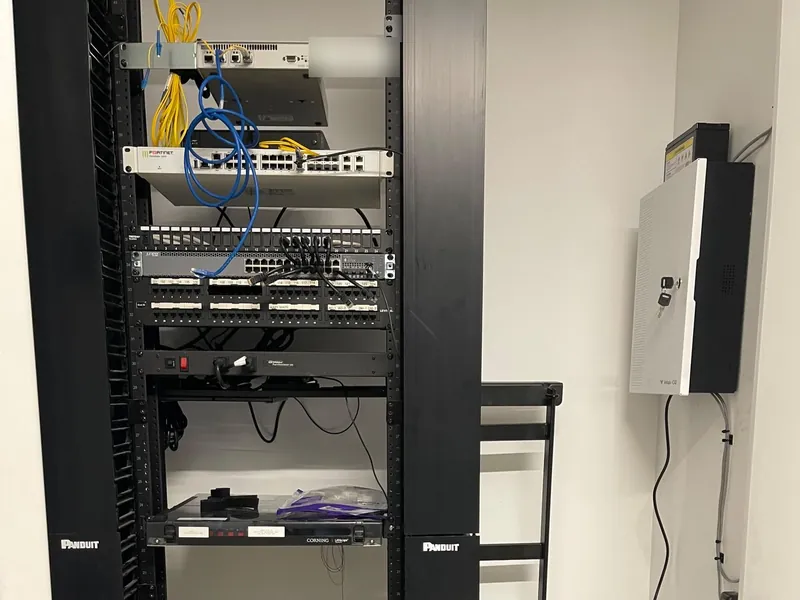 Network rack with a firewall, network switch and patch panel, beside a wall-mounted access control panel in the same equipment closet
