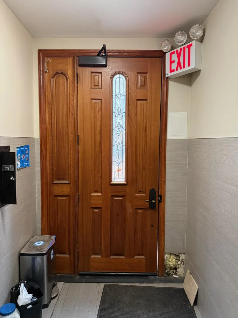 Wooden residential building entrance door with an overhead closer and an illuminated exit sign in a tiled vestibule