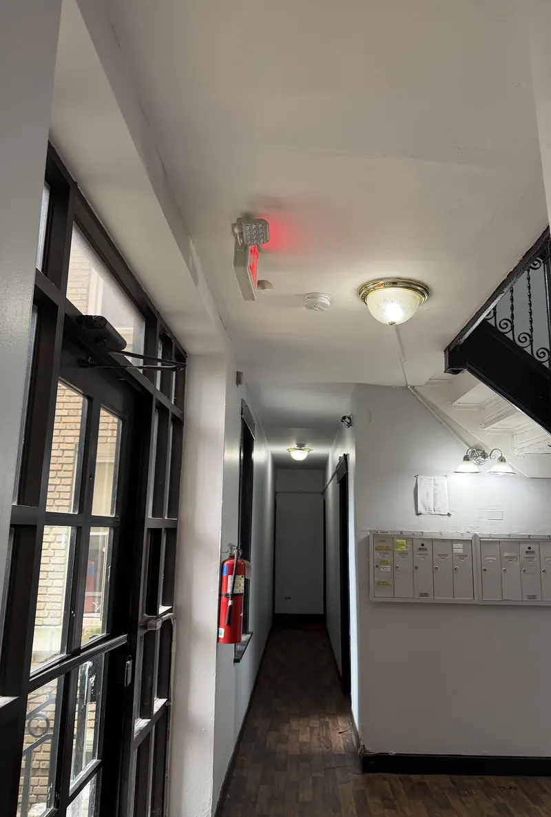 Residential building corridor with an illuminated exit sign, a fire extinguisher, mailboxes and an entrance door with an overhead closer