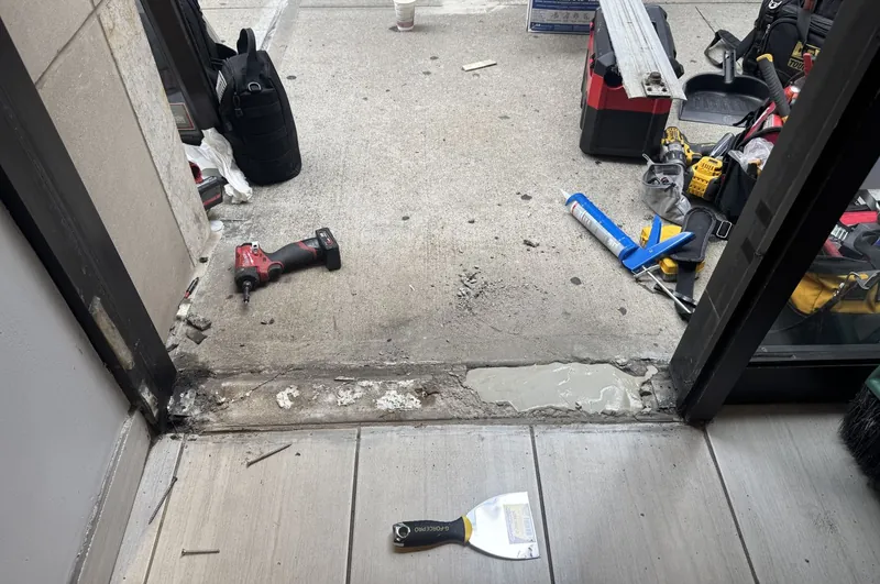 Storefront doorway with the door removed and the threshold area broken out for repair, with tools on the sidewalk